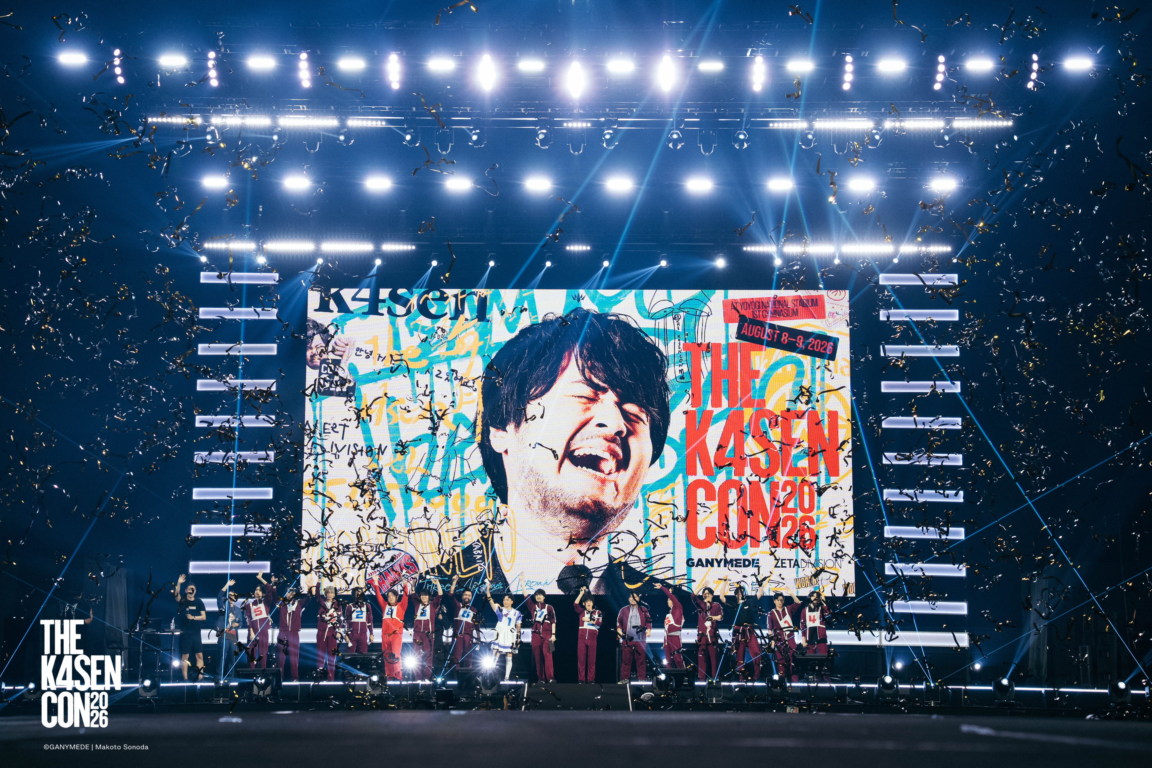 The k4sen Con 2026の大きなスクリーンと、ステージ上に並ぶ出演者の開催写真。